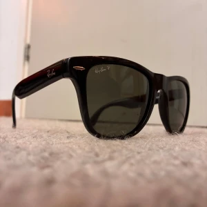 Ray-Ban Folding Wayfarer solglasögon - Snygga svarta Ray-Ban Folding Wayfarer solglasögon med klassisk wayfarer-form. De är hopfällbara och har mörka polariserade glas. Perfekta att ta med överallt tack vare den smidiga designen. Ikonisk logga på bågen och linsen.