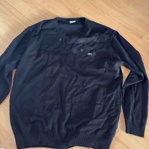 Svart stickad tröja från Lacoste XXL - Svart stickad tröja från Lacoste i storlek XXL med klassisk v-ringning och den ikoniska gröna krokodilloggan broderad på bröstet. Tröjan har långa ärmar och ribbade muddar vid ärmslut och nederkant. Perfekt för en clean och stilren look.