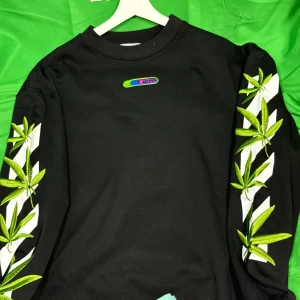 Svart off white tröja  - Svart sweatshirt från Off-White med stora vita grafiska tryck och broderade gröna blad på både ärmar och rygg. Tröjan har rund hals och lång ärm, perfekt för dig som vill sticka ut med en cool streetstyle-look.Liknande modell som originalet!