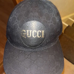 Svart Gucci keps med logga, storlek S - Svart keps från Gucci med klassiskt GG-mönster över hela kepsen och en rund läderpatch framtill med guldig GUCCI-text. Snygg och stilren modell som passar perfekt till streetwear. Kepsen är i storlek S (57 cm) och har en böjd skärm. Tar byten mot en svart eller beige kingsnake i S!