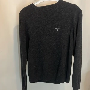 Mörkgrå stickad tröja från GANT - Mysig mörkgrå stickad tröja från GANT med rund halsringning och diskret logga broderad på bröstet. Perfekt för lager-på-lager och passar till allt. Klassisk och enkel design med långa ärmar och mjukt material. Kommer aldrig till användning men är väldigt fin på.