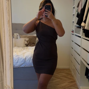 Brun one shoulder kortklänning - Säljer en brun kortklänning med one shoulder och cut out-detalj i sidan. Klänningen har en tight passform och är perfekt för dig som gillar en stilren men ändå edgy look. Materialet känns mjukt och följsamt mot kroppen. Superfin att styla med smycken och klackar.