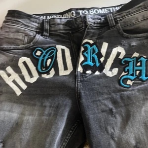 Svarta jeans från Hoodrich med tryck - Säljer ett par svarta jeans från Hoodrich med stor vit och blå text framtill. Jeansen har slitna detaljer, normal passform och klassisk femficksdesign. Snyggt Hoodrich-märke på bakfickan och bälte. Perfekta för dig som vill sticka ut. Jag är 185 och de passar perfekt, dock inte min stil riktigt!  Slim fit 28 w xs. 