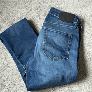 Mörkblå jeans från Jack&Jones  - Säljer ett par klassiska blåa jeans från Jack&Jones, i storlek 30x32 och modell Slim/Glenn. Jeansen har fem fickor och snygga kontrastsömmar. Jeansen har normal passform och raka ben, tillverkade i mjukt bomullstyg. Perfekta för dig som gillar stilrena och tidlösa jeans.