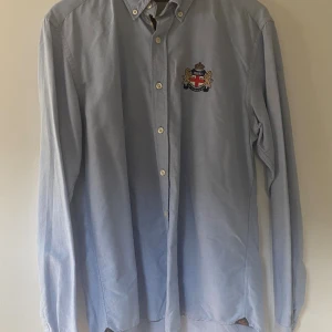 Ljusblå skjorta från Morris L - Klassisk ljusblå skjorta från Morris med broderad vapensköld på bröstet. Skjortan har button down-krage, vita knappar och lång ärm. Perfekt för dig som gillar stilrena och preppy detaljer.