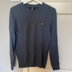 Blå Lyle & Scott stickad tröja - Snygg blå stickad tröja från Lyle & Scott med rund hals och klassisk gul logga på bröstet. Tröjan är tillverkad i 85% bomull och 15% ull, har ribbade muddar och lång ärm. Perfekt för dig som gillar stilren och enkel design.