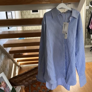 Blåvit randig skjorta från NA-KD - Oversized skjorta från NA-KD i blåvitt randigt mönster. Skjortan har klassisk krage, långa ärmar med breda manschetter och knappar framtill. Tillverkad i mjuk bomull för en chill och relaxed vibe.