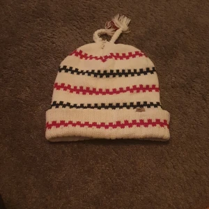 Randig vit mössa med tofsar - Säljer en vit stickad mössa med röda och svarta ränder. Mössan har uppvikt kant och två flätade tofsar på toppen. Tillverkad i 100% akryl, perfekt för kalla dagar. Liten logga på kanten.