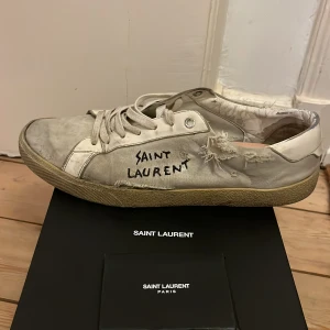 Saint Laurent vita sneakers med råa detaljer - Saint Laurent sneakers i vit canvas med handskriven logga på sidan. Skorna har slitna och trasiga detaljer för en edgy look, snörning framtill och beige gummisula. Perfekta för dig som gillar streetstyle och vill sticka ut. Allt og förutom sulorna dem är Loubs sulor 