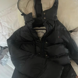 Svart pufferjacka Tommy Hilfiger Denim - Svart pufferjacka från Tommy Hilfiger Denim med hög krage, avtagbar huva och snörning i midjan. Jackan har en quiltad look, dragkedja och tryckknappar framtill samt diskret logga på bröstet. Fodret är ljust och mjukt, perfekt för kalla dagar.