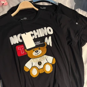 Svart Moschino t-shirt med nalle - Svart t-shirt från Love Moschino med broderad nalle och stora vita bokstäver framtill. T-shirten har rund hals, korta ärmar och coola detaljer i gult, vitt och rött. Perfekt statement-plagg för dig som gillar streetstyle och lekfull design. storlekar finns mellan S-XL