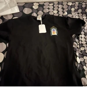 Svart t-shirt från Casablanca M - Svart t-shirt från Casablanca med färgglatt tryck av byggnad och blad på ryggen samt mindre tryck på bröstet. Klassisk rund hals och korta ärmar. Snygg och stilren design med coolt motiv. Levereras med originalpåse och tags kvar.