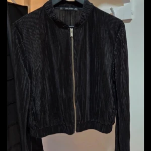 Svart plisserad bomberjacka Zara, M - Svart plisserad bomberjacka från Zara Basic med glansig finish. Jackan har dragkedja framtill, ribbad krage och muddar samt en croppad passform. Materialet är lätt och ger en cool textur. Perfekt för dig som gillar stilrena men unika plagg.