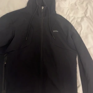 Svart hoodie från BOSS med dragkedja - Svart hoodie från BOSS med hel dragkedja framtill, huva med snörning och diskret BOSS-logga på bröstet. Tröjan har långa ärmar och praktiska fickor med dragkedja. Perfekt för en chill och stilren look.