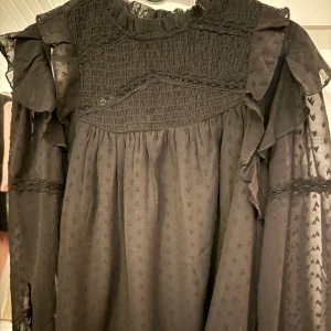 Svart volangkblus med spetsdetaljer - Supersnygg svart blus från gina med volanger på ärmarna, smock upptill och fina spetsdetaljer. Tunt, lätt material med diskret struktur och transparenta partier på ärmarna. Perfekt för dig som gillar romantisk och trendig stil.passar mig som är 164 bär vanligtvis xs-s så den är stor i storleken 