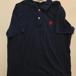 Mörkblå pikétröja från Timberland - Snygg marinblå pikétröja från Timberland med klassisk krage och två knappar framtill. Tröjan har ett rött broderat Timberland-logo på bröstet och är tillverkad i mjuk bomull. Perfekt för en stilren och avslappnad look.                                                         STORLEK M