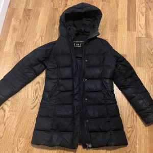 Svart dunjacka med huva från Boomerang - Säljer en svart, lång dunjacka från Boomerang modell SIRI. Jackan har huva, två dragkedjeförsedda fickor och quiltad design. Perfekt för kalla dagar och har en stilren look med raka linjer och diskret logga på insidan. B