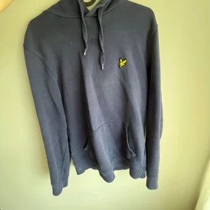 Mörkblå hoodie från Lyle & Scott - Mörkblå hoodie från Lyle & Scott med klassisk gul logga på bröstet. Tröjan har huva med snörning, känguruficka och långa ärmar. Perfekt för chill dagar och enkel att matcha med jeans eller joggers.