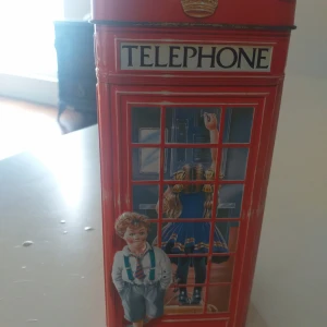 Röd telefonkiosk sparbössa - Dekorativ sparbössa i form av en klassisk brittisk telefonkiosk, tillverkad i plåt med färgglada motiv av barn. Sparbössan har ett myntinkast på toppen och är i gott skick med endast mindre tecken på användning. Perfekt som present eller inredningsdetalj.