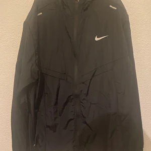 Svart Nike miler  - Svart vindjacka från Nike med klassisk vit Swoosh-logga på bröstet. Jackan har hel dragkedja, huva och reflekterande detaljer på axlarna. Tillverkad i lätt polyester, perfekt för blåsiga dagar. Sportig och stilren design som funkar till streetwear.