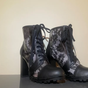 Svarta spets stövletter med klack - Säljer ett par svarta stövletter med hög klack och snörning. Skorna har transparenta partier med blommig spets och detaljer i syntetläder. Grov platt sula och rund tå ger en edgy vibe. Perfekta för dig som vill sticka ut med coola detaljer och unik design.