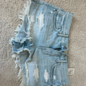 Ljusblå slitna jeansshorts Abercrombie - Säljer ett par ljusblå jeansshorts från Abercrombie & Fitch med slitna detaljer och fransiga kanter. Klassisk femficksmodell med dragkedja och knapp. Perfekta för sommaren och ger en avslappnad vibe.