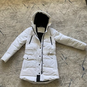 Vit pufferjacka från Samsa Collection XS - HELT NY OANVÄNT !!!Vit lång pufferjacka från Samsa Collection i storlek XS. Jackan har huva, svarta dragkedjor, tryckknappar och stora fickor framtill. Insidan är svart och jackan är quiltad för extra värme. Perfekt för kalla dagar och har en clean, modern look.