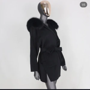 Svart kappa med fuskpäls och bälte - Stilren svart kappa med stor fuskpäls runt kragen och knytskärp i midjan. Jackan har lång ärm, omlottmodell och slits i sidorna. Perfekt för dig som vill ha en elegant och trendig look. Helt okej skick inte använd mycket egna bilder kan fås.