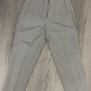 Grå kostymbyxor från Zara - Snygga grå kostymbyxor från Zara med pressveck och hög midja. Byxorna har raka ben och diskreta sidfickor, perfekta för en clean och stilren look. Materialet är mjukt och känns lyxigt mot huden.