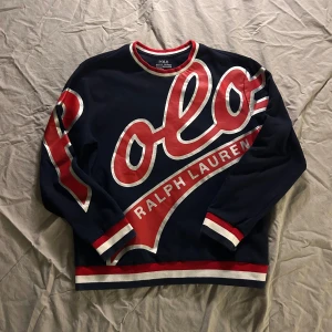 Mörkblå tröja Polo Ralph Lauren - Snygg mörkblå tröja från Polo Ralph Lauren med stor röd och vit logga över hela framsidan och ena ärmen. Tröjan har ribbade muddar och krage i rött, vitt och blått. Tillverkad i bomull och polyester, perfekt för dig som gillar sportig och trendig stil. Storleken är en aning minskad och är mer än Large/medium än en large/XL. Skickas samma dag och pris kan diskuteras vid snabb affär!