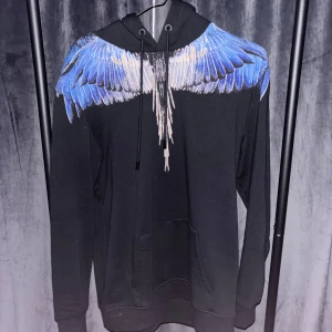 Marcelo burlon icon wings  - Svart hoodie med huva och dragsko, prydd med ett stort blått och vitt vingmönster över axlar och bröst. Tröjan har känguruficka framtill och långa ärmar. Perfekt för dig som vill sticka ut med en unik och cool design.