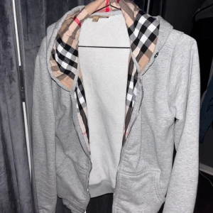 Grå hoodie med Burberry-rutor - Grå hoodie från Burberry med dragkedja och klassiskt rutigt foder i huvan. Tröjan har långa ärmar, två fickor framtill och ribbade muddar. Perfekt för dig som vill ha en stilren men ändå lyxig look.