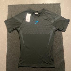 Gymshark onyx compressionshirt - Snygg svart t-shirt från Gymshark i funktionsmaterial, perfekt för träning. T-shirten har korta ärmar, diskret mönster i grått och en blå Gymshark-logga på bröstet. Slimmad passform och sportig look.