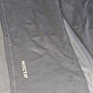 Svarta NOCTA x Nike mjukisbyxor - Svarta mjukisbyxor från NOCTA x Nike med guldfärgade detaljer och loggor. Byxorna har snörning i midjan, dragkedjeficka på sidan och elastisk midja. Tillverkade i mjukt material som passar perfekt för chill eller träning.