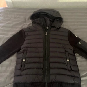 Svart pufferjacka från Moncler - Säljer en svart pufferjacka från Moncler med stickade ärmar och huva. Jackan har dragkedja framtill, två fickor med tryckknappar och en extra ficka med dragkedja på ärmen. Klassisk Moncler-logga på vänster ärm. Perfekt för kyliga dagar.