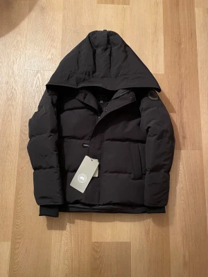 Svart pufferjacka från Canada Goose - Svart pufferjacka från Canada Goose med stor huva och diskret logotyp på ärmen. Jackan har quiltad design, dragkedja och knappar framtill samt ribbade muddar vid ärmslut. Perfekt för kalla dagar och har en clean, modern look.