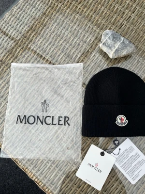 Svart Moncler mössa  - Svart ribbstickad mössa från Moncler med klassisk uppvikt kant och ikonisk Moncler-logga framtill. Mössan är tillverkad i mjuk ull och har en stilren look som passar perfekt till kalla dagar. Medföljer originalpåse och taggar.https://code.moncler.com/?QR=35225294UPUY