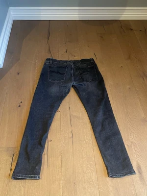 Svarta jeans från Nudie Jeans, W30 L28 - Säljer ett par svarta jeans från Nudie Jeans med klassisk femficksdesign och snygga detaljer på bakfickorna. Jeansen har en smal passform och är tillverkade i mjukt bomullsmaterial. Perfekt för dig som gillar stilrena och tidlösa jeans.
