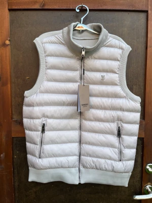 Vit dunväst från Burberry XL - Snygg vit dunväst från Burberry med quiltad design och ribbade detaljer vid krage, ärm och nederkant. Västen har två fickor med dragkedja framtill och en innerficka med zip. Perfekt för lager-på-lager och har en stilren look med diskret Burberry-logga på bröstet.