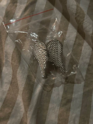 Spännknäppen - Säljer ett par silvriga spännknäppen till byxor formade som vingar med ett randigt mönster. Man ska sätta de i midjan om byxorna sitter löst💕