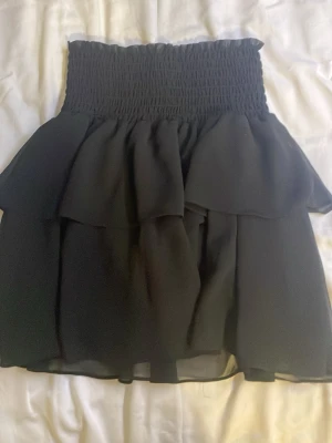 Svart volangkjol från Chelsea - Säljer en svart kortkjol från Chelsea med bred smockad midja och dubbla volanger. Kjolen är gjord i ett lätt och flödande syntetmaterial som ger en snygg siluett. Perfekt för dig som gillar trendiga och feminina plagg med extra volym.