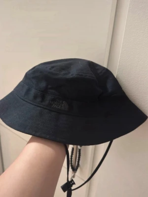 Svart bucket hat från The North Face - Säljer en stilren svart bucket hat från The North Face i storlek S-M. Hatten är tillverkad i 100% bomull och har ett justerbart hakband för extra säkerhet. Perfekt för att lyfta din streetwear-look och skydda mot solen.