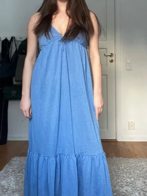 Blå långklänning med volang - Säljer en blå långklänning med v-ringad hals och smala axelband. Klänningen har en luftig passform och en bred volang längst ner som ger extra rörelse. Perfekt för varma dagar och har en enkel, stilren look.