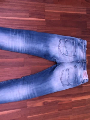 Slim Glenn ljusblå slitna jeans - Säljer ett par Slim Glenn jeans från Jack & Jones i ljusblå tvätt med slitningar och hål på framsidan. Jeansen har klassisk femficksdesign, smal passform och coola detaljer på insidan av midjan. Perfekt för dig som gillar en avslappnad streetstil.