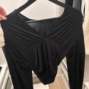 Svart omlottopp från NLY ONE - Säljer en svart omlottopp från NLY ONE i storlek XS. Toppen har lång ärm, v-ringning och snygga draperade detaljer framtill som ger en cool look. Materialet är mjukt och stretchigt, perfekt att styla med jeans eller kjol.