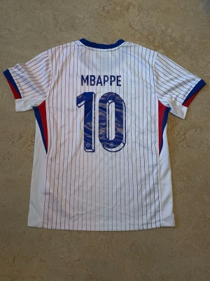 Nike Frankrike Mbappé tröja #10 - Säljer en vit Nike Frankrike landslagströja med blå och röda ränder, nummer 10 och Mbappé på ryggen. Tröjan har blå detaljer vid krage och ärmslut samt landslagets tupp-logga på bröstet. Tillverkad i polyester. Aldrig använt    PRIS KAN DISKUTERAS!!