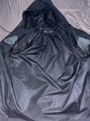 Svart vindjacka från Under Armour - Svart vindjacka från Under Armour med huva och gråa detaljer på ärmarna. Jackan har elastiska muddar vid ärmslut och en stor logga på vänster ärm. Perfekt för blåsiga dagar och sportiga aktiviteter. Snygg och funktionell design.