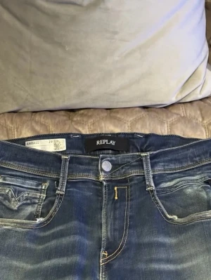 Replay Anbass jeans blå denim  - Säljer ett par Replay Anbass jeans i klassisk blå tvätt med snygga slitningar och lätt distressed detalj på ena benet. Modellen har normal passform, fem fickor och gul kontrastsöm. Perfekt för dig som gillar en cool och avslappnad stil.