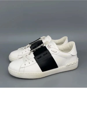 Valentino Garavani Open sneakers - Säljer ett par snygga Valentino Garavani Open sneakers i vitt skinn med bred svart läderdetalj över snörningen. Klassisk rund tå och platt sula, med coola nitar på hälen. Perfekt för dig som gillar stilrena och lyxiga sneakers.
