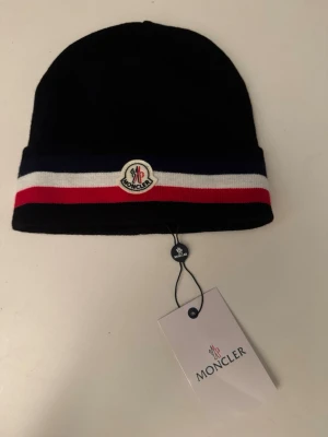 Moncler mössa med ränder - Snygg svart mössa från Moncler med breda ränder i marinblått, vitt och rött längs kanten. Mössan har Moncler-logga framtill och är stickad i mjukt material. Perfekt för dig som vill ha en clean och stilren look med lite färg.